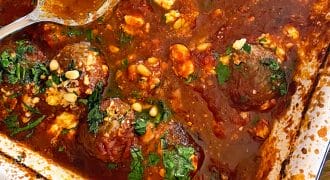 A baking dish filled with meatballs in a rich tomato sauce, topped with crumbled cheese, chopped herbs, and pine nuts. Two metal serving spoons rest in the dish.