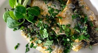 Grilled fish fillets garnished with chopped herbs, served with a wedge of lemon and a small side of fresh salad leaves on a white plate.