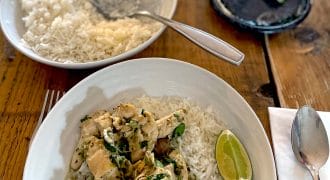 A bowl of white rice topped with chicken, vegetables, and herbs sits on a wooden table next to a lime wedge, spoon, serviette, plate with another lime wedge, and a bowl of plain rice with a spoon.
