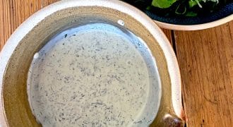 A rustic ceramic bowl filled with creamy herb dressing sits on a wooden table next to a dark bowl of fresh rocket and cucumber salad.