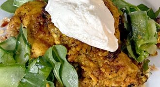 Seasoned roasted chicken topped with a dollop of creamy white sauce, served on a bed of rocket leaves and cucumber ribbons, garnished with chopped nuts on a white plate.