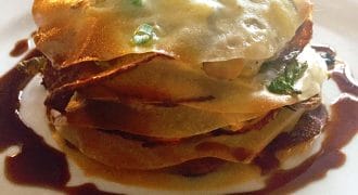 A plated dish featuring a layered savoury pastry with a golden-brown top, garnished with chopped spring onions and surrounded by a dark sauce on a white plate.