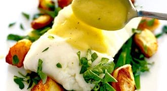 A spoon drizzles sauce over a piece of white fish on a bed of green beans and roast potatoes, garnished with fresh chopped herbs, served on a white plate.