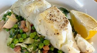 A white plate with two pieces of seasoned, cooked white fish on a bed of green peas, leafy greens, and diced vegetables, garnished with a lemon wedge and served with a light sauce.