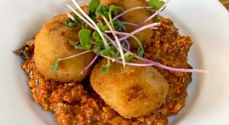 Three golden brown, breadcrumbed croquettes served on a bed of chunky red sauce, garnished with fresh green microgreens, on a white plate set on a wooden table.