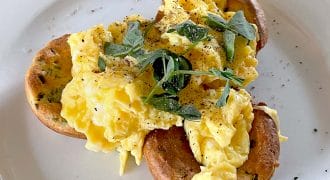 Scrambled eggs served on two toasted crumpets, garnished with fresh herbs and cracked black pepper, on a white plate.