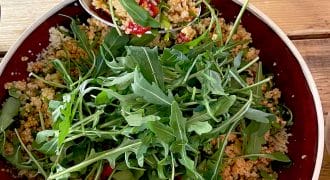 A bowl of couscous salad with chopped vegetables, garnished with fresh rocket leaves, sits on a wooden table. A spoon is serving a portion of the salad from the bowl.
