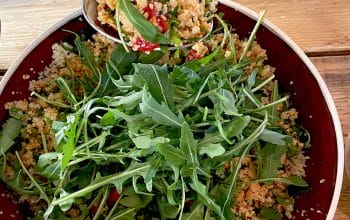 A bowl of couscous salad with chopped vegetables, garnished with fresh rocket leaves, sits on a wooden table. A spoon is serving a portion of the salad from the bowl.