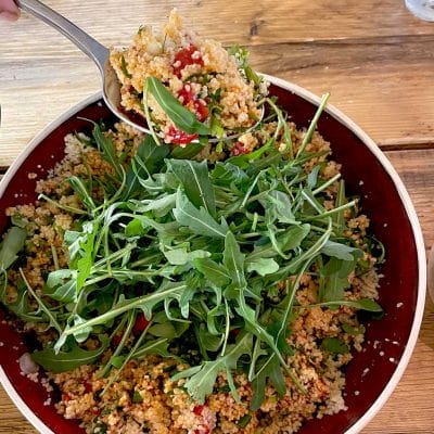 A bowl of couscous salad with chopped vegetables, garnished with fresh rocket leaves, sits on a wooden table. A spoon is serving a portion of the salad from the bowl.