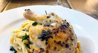 A roasted chicken leg, seasoned with herbs, is served on a bed of fettuccine pasta with greens on a white plate. In the background, a tray with more chicken and a glass of wine can be seen.
