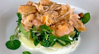 A plated dish featuring flaked salmon on a bed of fresh greens and sliced potatoes, served atop a layer of creamy white sauce on a white plate.