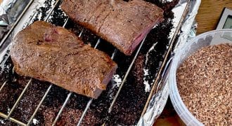 Two pieces of cooked meat rest on a metal rack inside a foil-lined roasting tin. Beside the tin is a plastic container filled with wood chips, likely for smoking the meat.