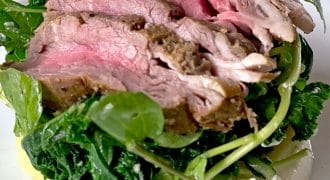 Slices of medium-rare roast beef are served on a bed of leafy greens and watercress atop sliced potatoes with a creamy white sauce, arranged on a white plate.