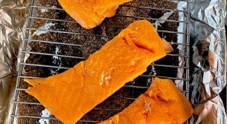 Three pieces of raw salmon fillet are placed on a metal rack over a layer of brown wood chips on aluminium foil, ready to be smoked or cooked.