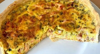 A baked quiche with a golden crust and a savoury filling, featuring visible pieces of vegetables and meat, sits on a white plate with a large slice removed.