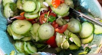 A bowl of fresh cucumber salad with sliced cucumbers, cherry tomatoes, onions, and coriander, served with metal tongs on a wooden table.