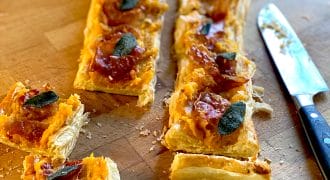 Rectangular puff pastry tart topped with orange mashed squash, crispy sage leaves, and slices of cured meat, cut into pieces on a wooden surface next to a knife.