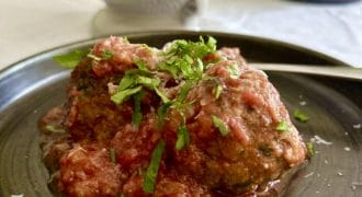 Two meatballs topped with tomato sauce and chopped fresh herbs are served on a dark plate. Some grated cheese is visible on top and around the plate, with a spoon resting beside the meatballs.
