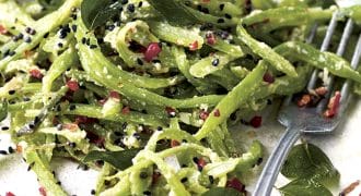 A plate of green pasta topped with black seeds, red chilli flakes, grated cheese, and fresh herbs. A fork rests on the plate beside the pasta.