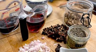 Chopped shallots, minced garlic, dried mushrooms, dried herbs, red wine, and cream are arranged on a wooden kitchen worktop, ready for cooking. Measuring jugs and glass jars hold some of the ingredients.