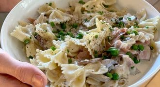A person holds a white bowl filled with farfalle pasta in a creamy sauce, mixed with peas, ham, mushrooms, and garnished with chopped chives and ground black pepper.