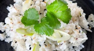 A close-up of a serving of seasoned white rice garnished with coriander leaves, sliced spring onions, and a sprinkle of black sesame seeds, presented on a dark plate.