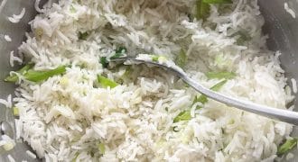 A pot of cooked white basmati rice mixed with chopped spring onions, with a metal spoon resting inside the pot.