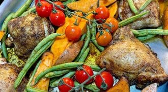 Baked chicken thighs, sweet potato wedges, green beans, and vine tomatoes arranged on a metal tray, seasoned and ready to eat.