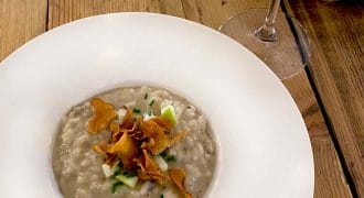 A white bowl of creamy risotto topped with crispy crisps and chopped herbs sits on a wooden table next to a glass of rosé wine.