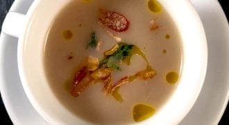 A white cup on a white saucer filled with creamy soup, garnished with crispy fried shallots, green herbs, and drops of oil, set on a dark surface.