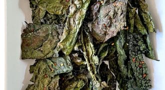A white rectangular dish filled with crispy, baked kale crisps. The leafy crisps are dark green and slightly curled, with visible seasoning flakes scattered on their surfaces.