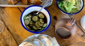 A wooden table set with a tray of breaded chicken goujons, a bowl of sliced cucumbers in sauce, a bowl of shredded lettuce, a pile of folded flatbreads, and a brown pottery jug.