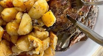 A white plate holding roast potatoes seasoned with herbs and a piece of roast meat with a fork and knife cutting into it, on a wooden table.