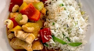 A white bowl filled with white rice mixed with vegetables, served alongside stir-fried chicken with red and yellow peppers, celery, pineapple, and topped with cashew nuts in a savoury sauce.