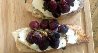 Three slices of crusty bread topped with creamy cheese, roasted grapes, pine nuts, and fresh herbs are arranged on a wooden serving board.