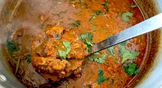 A pot of rich, red-brown curry garnished with fresh coriander, with a ladle lifting a portion of the meat and sauce. Steam rises from the hot dish, indicating it is freshly cooked.