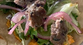 A close-up of a flatbread topped with mixed leaves, sliced red onions, grilled meat pieces, and an orange spread, ready to be wrapped.