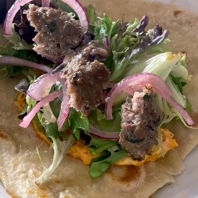 A close-up of a flatbread topped with mixed leaves, sliced red onions, grilled meat pieces, and an orange spread, ready to be wrapped.