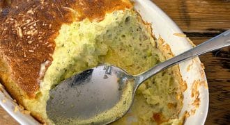 A close-up of a baked courgette soufflé in a white oval dish, with a spoon resting inside and part of the soufflé scooped out, showing its fluffy, airy texture. The dish sits on a rustic wooden table.