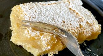 A slice of lemon bar covered with icing sugar sits on a dark plate, with a fork cutting into its soft, creamy texture.