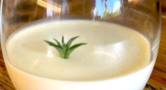 A round glass filled with a creamy white dessert, topped with a small green herb sprig, sits on a wooden table. The background is softly reflected on the glass.