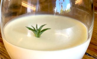 A round glass filled with a creamy white dessert, topped with a small green herb sprig, sits on a wooden table. The background is softly reflected on the glass.