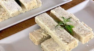Rectangular pieces of shortbread are neatly stacked on two white rectangular plates, dusted with icing sugar and garnished with a small sprig of green herb. The plates rest on a wooden surface.