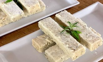 Rectangular pieces of shortbread are neatly stacked on two white rectangular plates, dusted with icing sugar and garnished with a small sprig of green herb. The plates rest on a wooden surface.