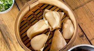 A bamboo steamer with five folded bao buns sits on a wooden table, surrounded by bowls of spring onions, herbs, a jar of sauce, tongs, and a pot of shredded meat.