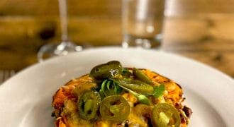 A baked sweet potato half topped with melted cheese, beans, and sliced jalapeños sits on a white plate. In the background are a glass of red drink and a tall glass of water on a wooden table.
