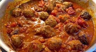 A frying pan filled with meatballs simmering in a rich tomato sauce with chunks of tomatoes, set on a hob with tiled splashback in the background.