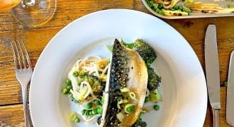 A white plate with grilled fish on top of noodles, broccoli, peas, and bean sprouts. A fork and knife are on either side, with a glass of drink and a bottle of soy sauce on a wooden table.