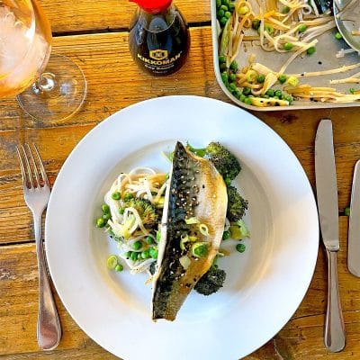 A white plate with grilled fish on top of noodles, broccoli, peas, and bean sprouts. A fork and knife are on either side, with a glass of drink and a bottle of soy sauce on a wooden table.