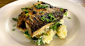 Grilled fish fillet garnished with chopped chives, served on a bed of salad leaves and creamy potato salad, presented on a white plate atop a wooden table.